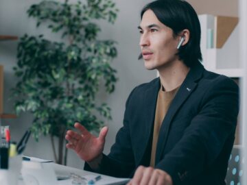 A focused man works in a modern office environment wearing earbuds, seemingly engaged in a video call.