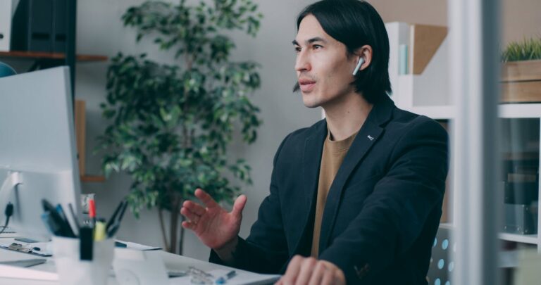 A focused man works in a modern office environment wearing earbuds, seemingly engaged in a video call.