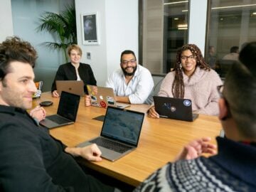 four people all on laptops, two men and two women, listen to person talking in a board meeting